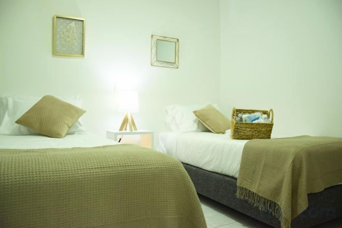 Une chambre avec deux lits, une table de chevet avec une lampe et un panier posé sur l’un des lits.