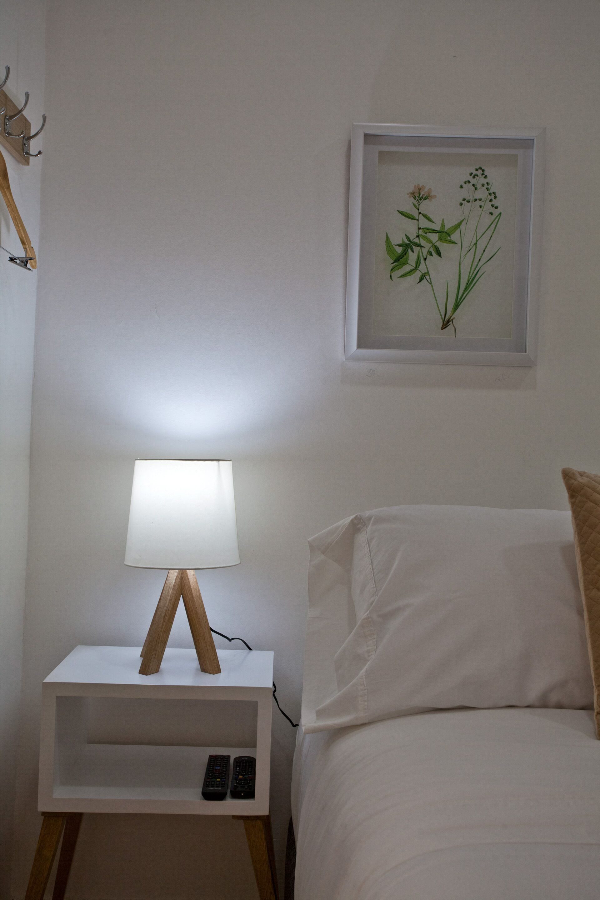 A neatly arranged bedroom with a bedside table, a lamp, a framed botanical print, and a bed with pillows.