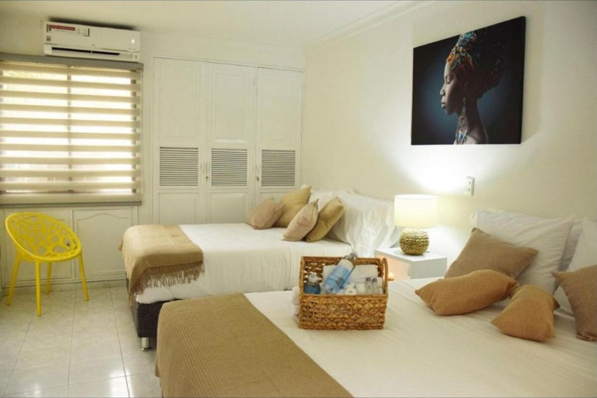 Une chambre d’hôtel avec deux lits, une chaise jaune, un climatiseur fixé au mur, un tableau encadré et une table de chevet avec une lampe.