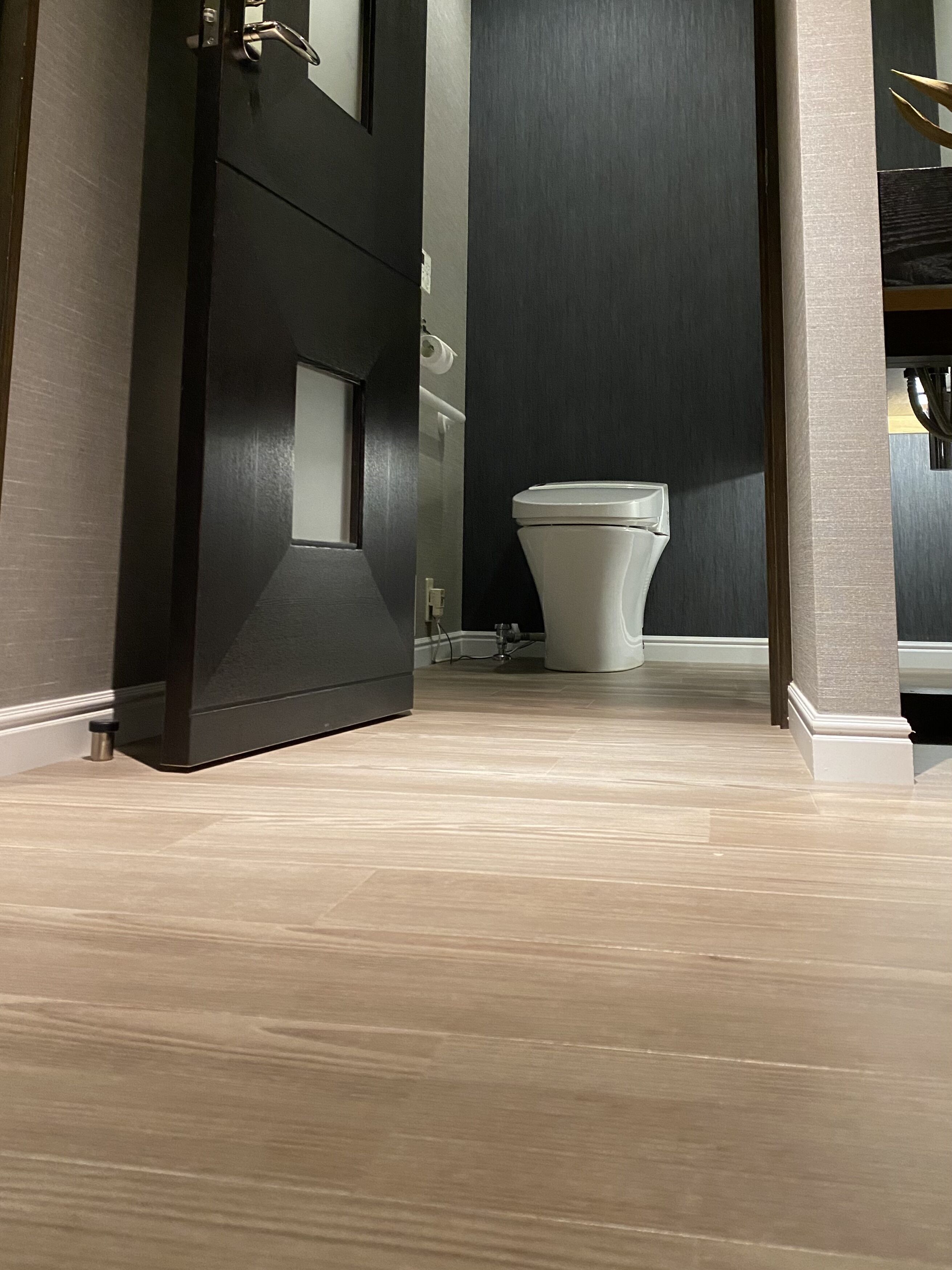 Un baño moderno con una puerta oscura, un inodoro blanco y un suelo de madera clara.
