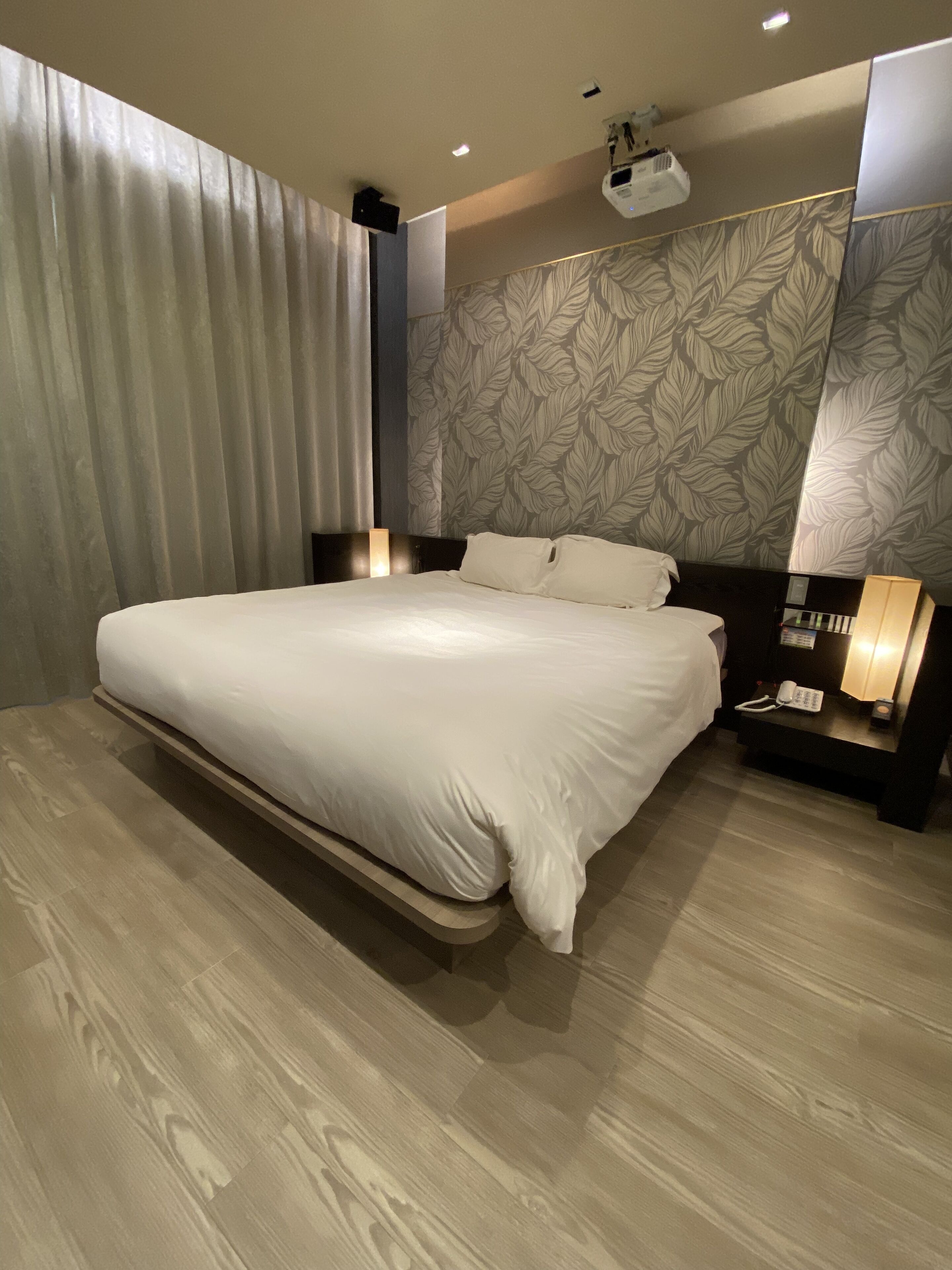 A modern hotel room with a large bed, a nightstand with a lamp, and a wall-mounted projector.