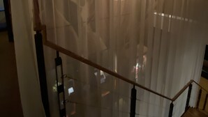Un escalier avec une rampe en bois et une balustrade en verre, menant à une pièce dotée d’une grande fenêtre recouverte de rideaux légers.