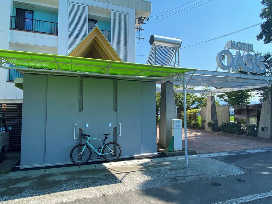En hotelindgang med et overdækket venteområde, en cykel parkeret indeni og et skilt med hotellets navn.