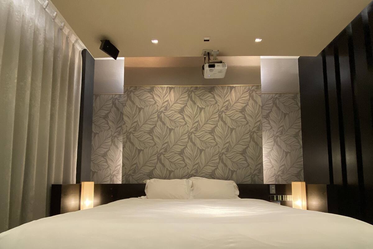 A modern hotel room with a large bed, a ceiling light, and a wall-mounted projector.