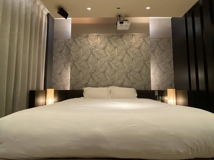 A modern hotel room with a large bed, a ceiling light, and a wall-mounted projector.