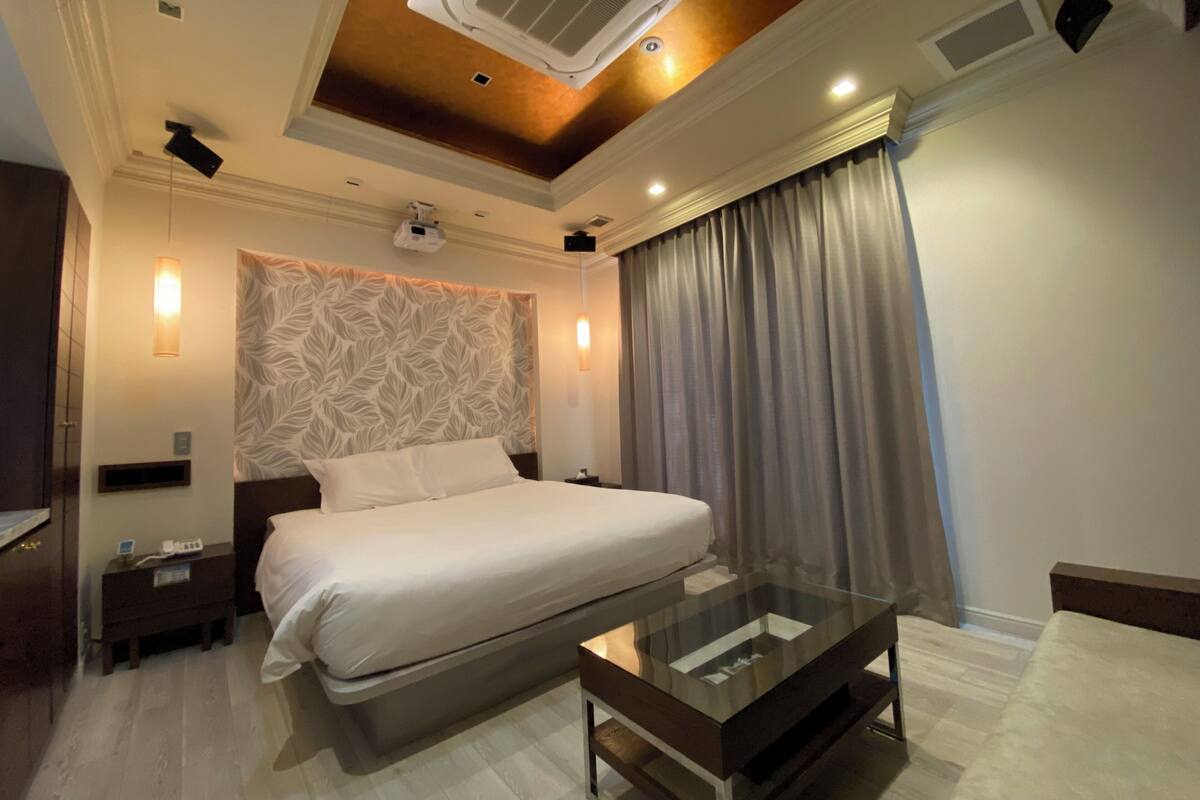 A modern hotel room with a large bed, a glass coffee table, a ceiling fan, and a large window with curtains.