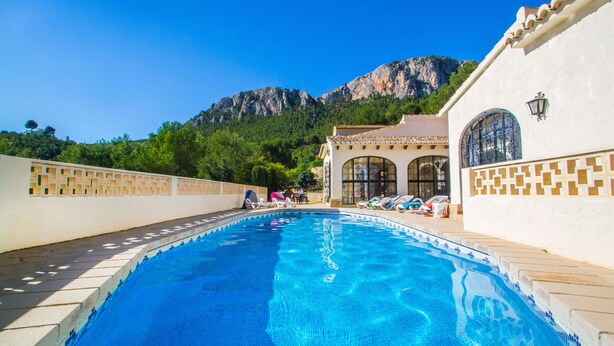 A swimming pool with lounge chairs and a building with arched windows, surrounded by greenery and mountains. - Villa Moskoa - PlusHolidays (Calpe)