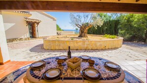 An outdoor dining area with a round table set for six, surrounded by stone walls and a view of the sea. - Villa Moskoa - PlusHolidays (Calpe)