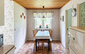 A kitchen with a dining table, chairs, a hanging light fixture, a window with curtains, and a clock on the wall.