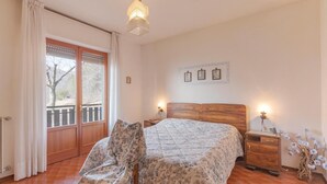 Un dormitorio con una cama, una silla, una mesita de noche, una lámpara y un ventanal con cortinas.
