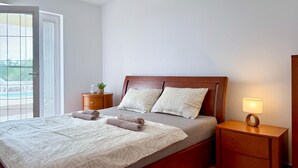 Ein Schlafzimmer mit einem Bett, zwei Kissen, einem Nachttisch mit Lampe, einem Beistelltisch und einem Fenster mit Jalousien.