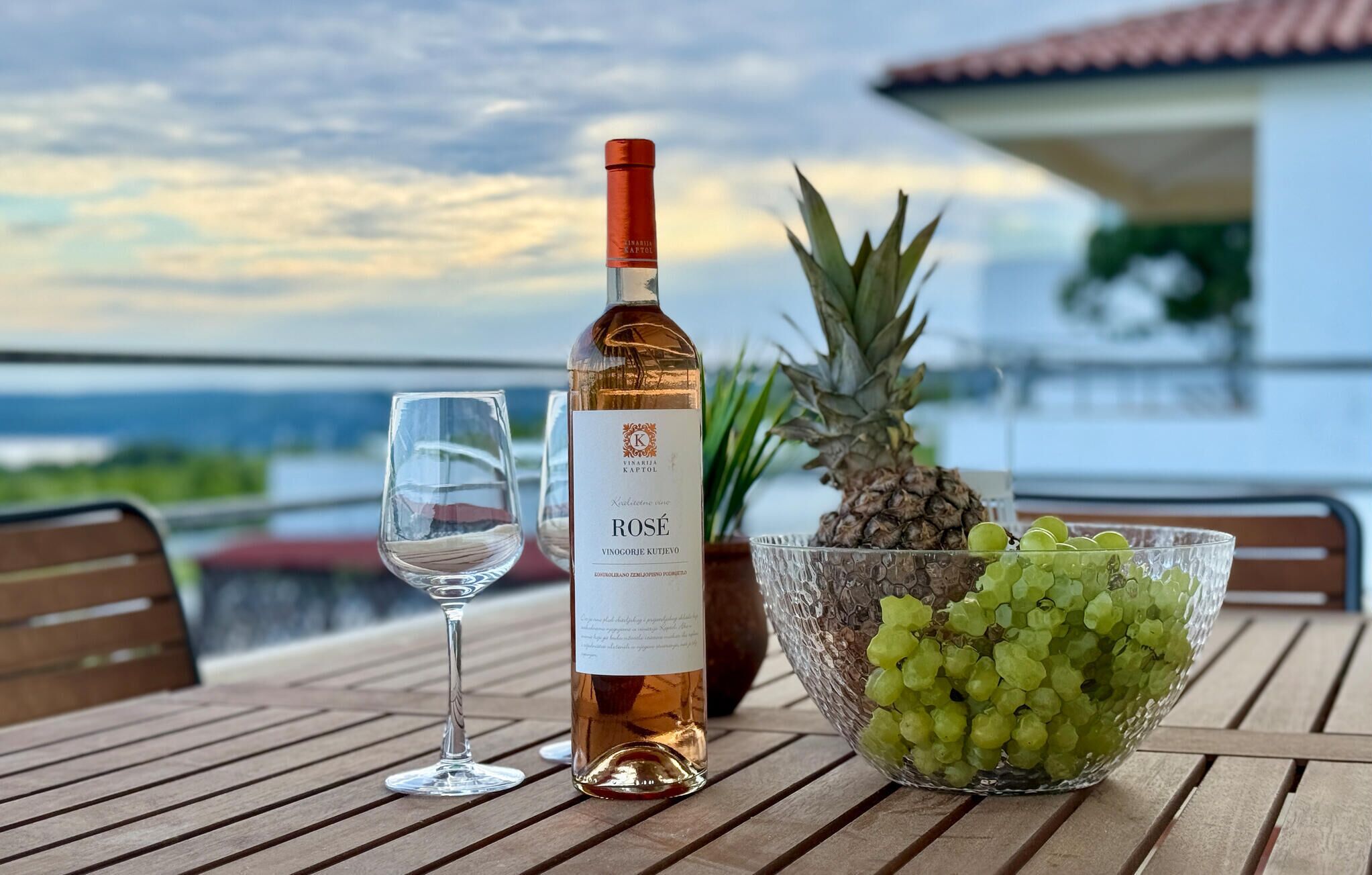 En flaske rosévin, et glas, en ananas og en skål med druer på et træbord med udsigt til havet.