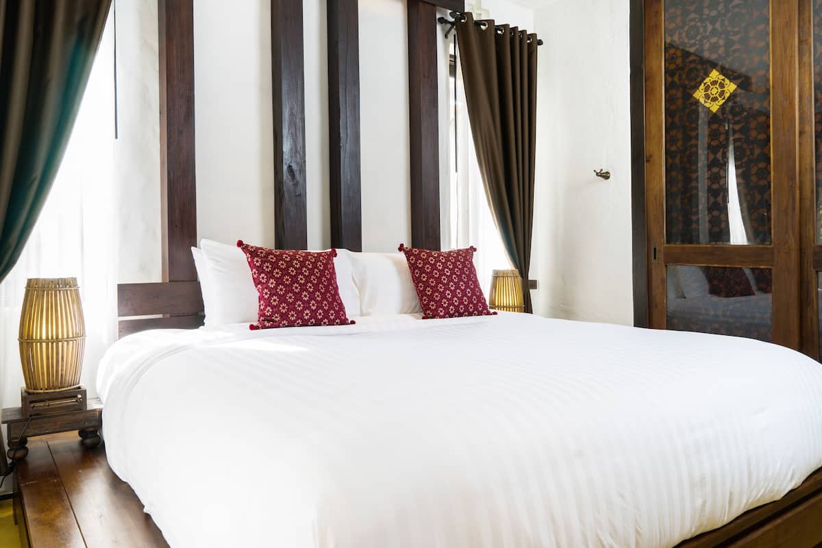 A large bed with white bedding and red patterned pillows, wooden headboard, and a view of a traditional building through a window with curtains.