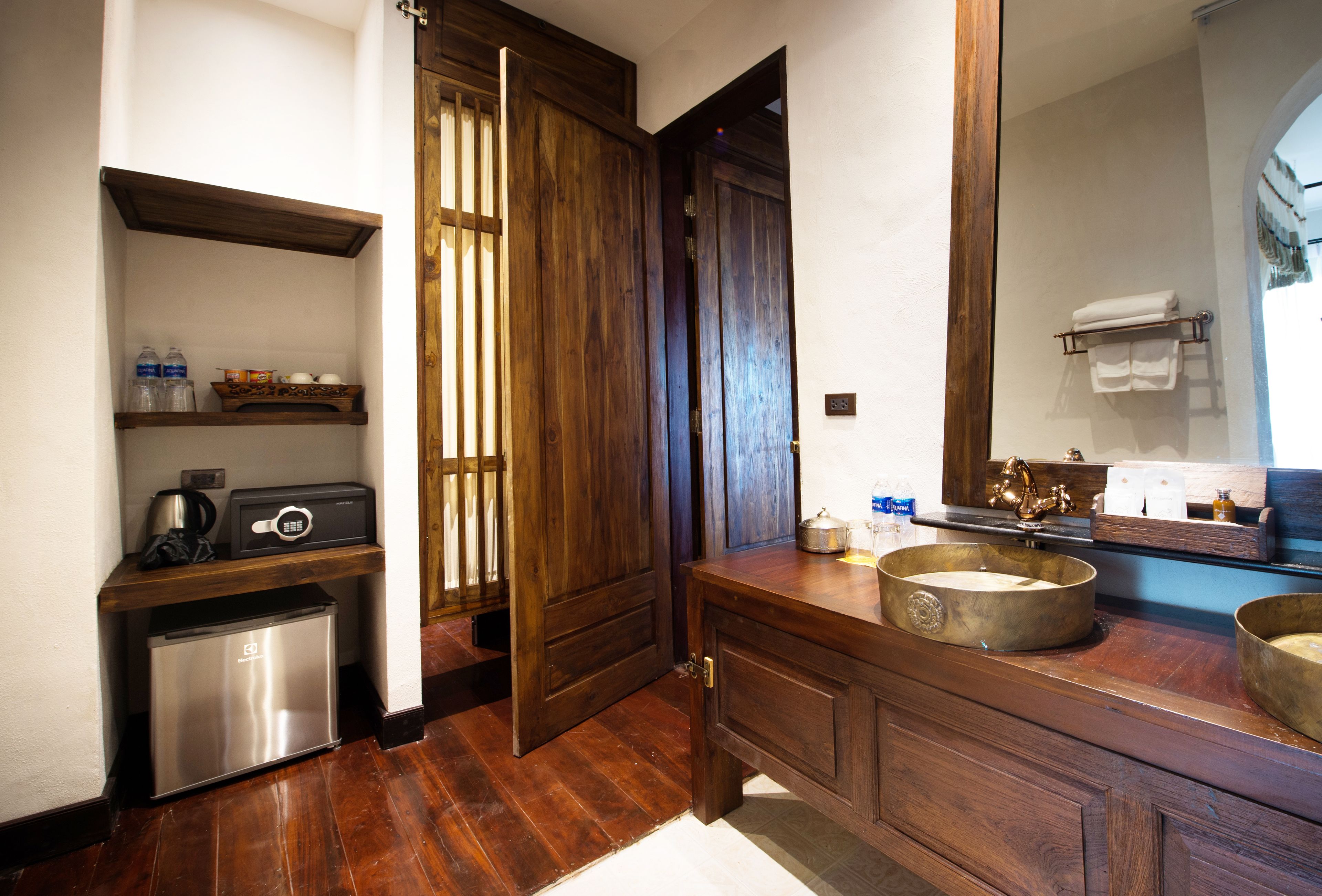 A compact bathroom with a wooden vanity, a sink, and a mirror. There is a small refrigerator and a shelf with toiletries.