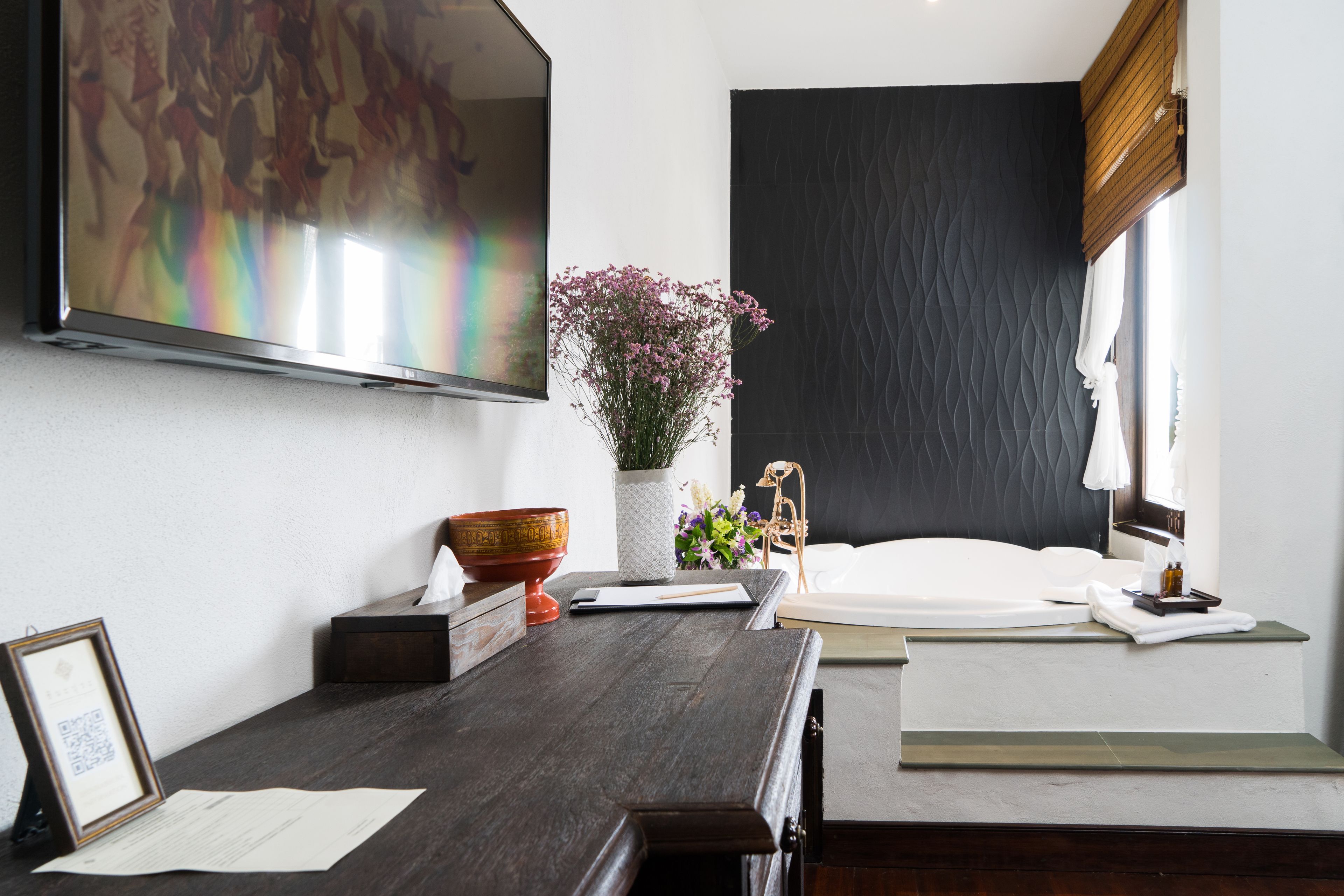Una habitación de hotel moderna con un televisor de pantalla plana grande, un escritorio de madera con un florero y una cama con cabecera oscura, y una ventana con vistas al exterior.