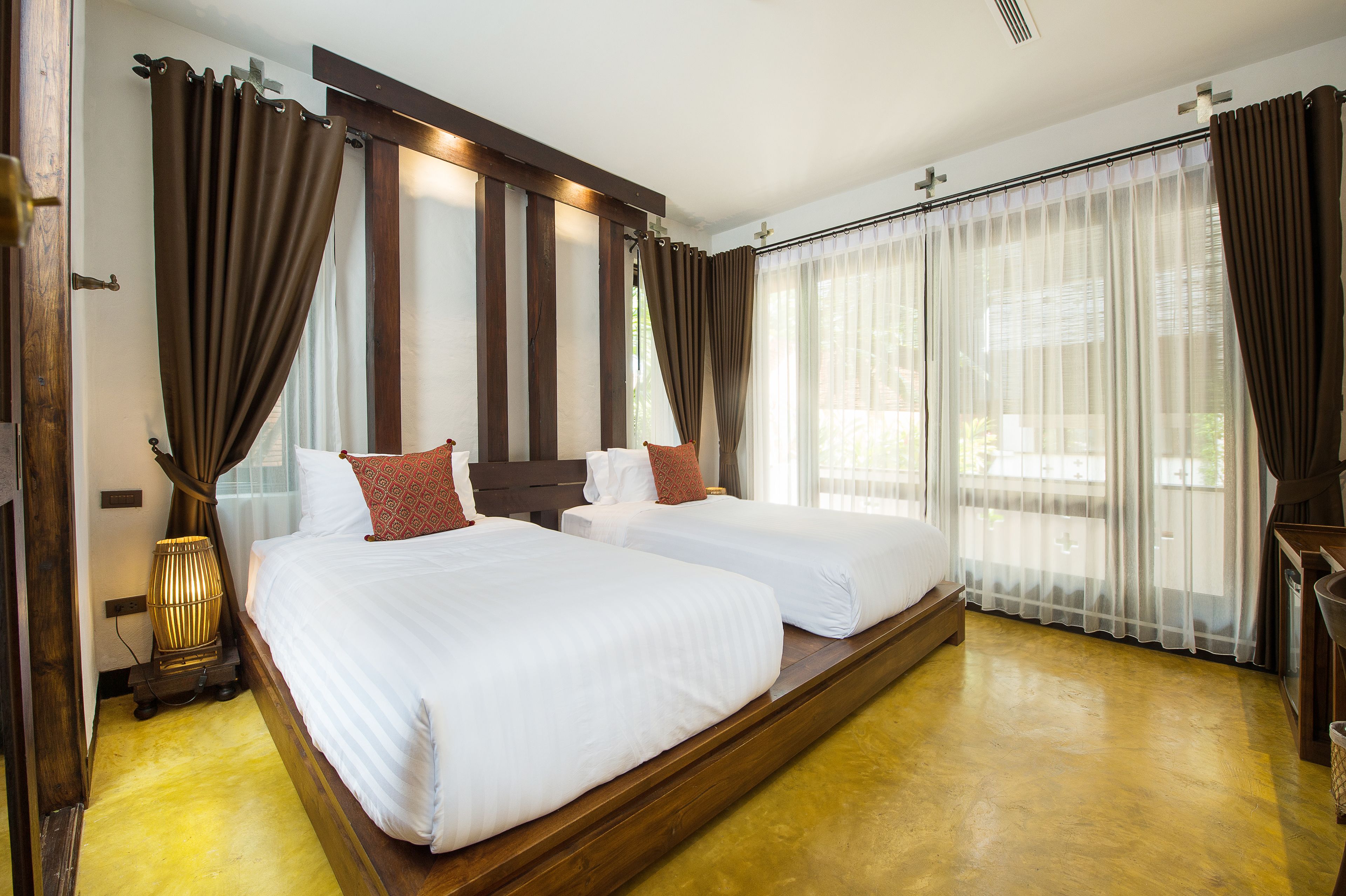 A hotel room with two beds, wooden flooring, and large windows with curtains.
