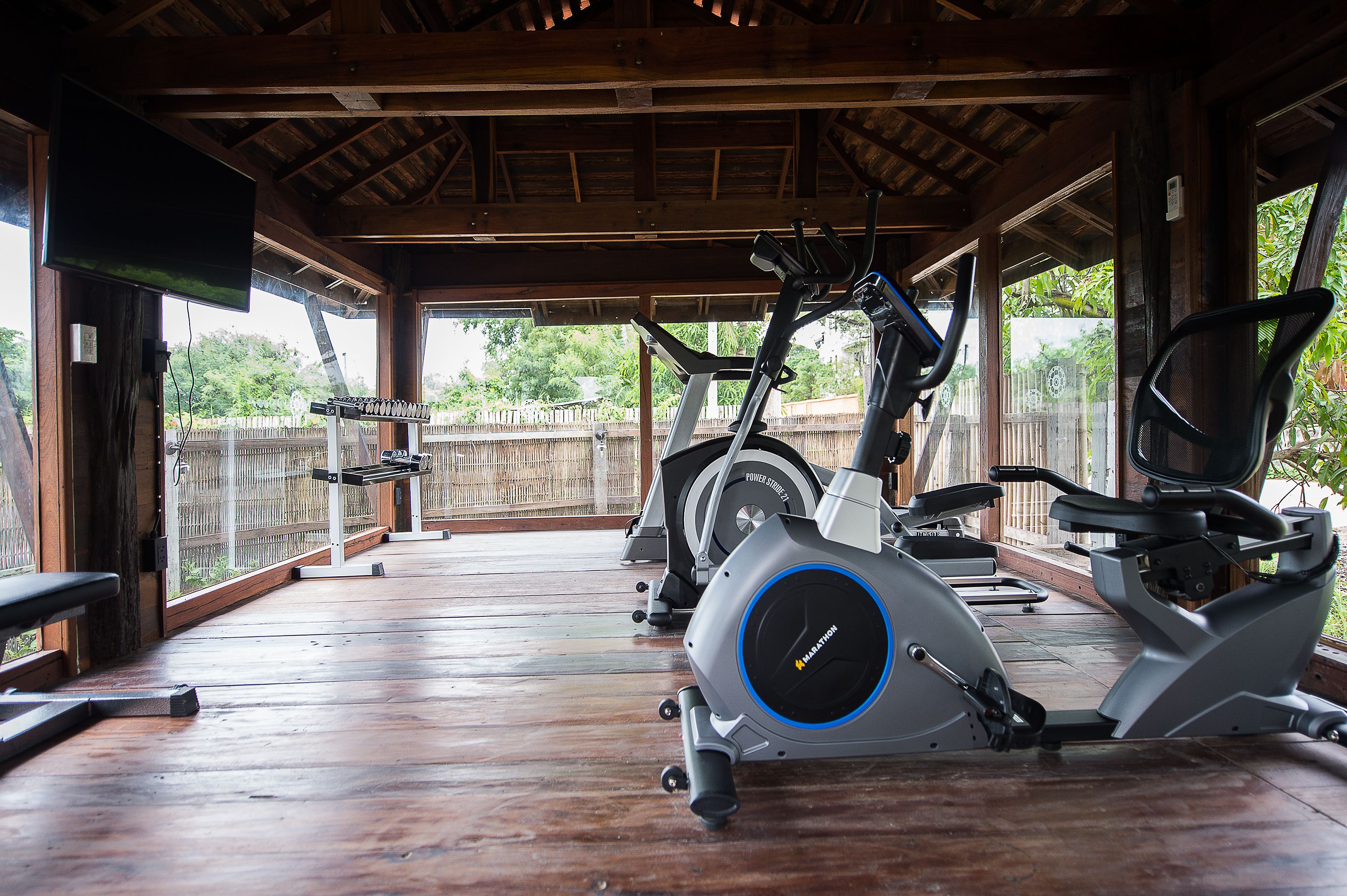 A covered outdoor gym area with exercise equipment, wooden flooring, and a view of a fence and greenery.