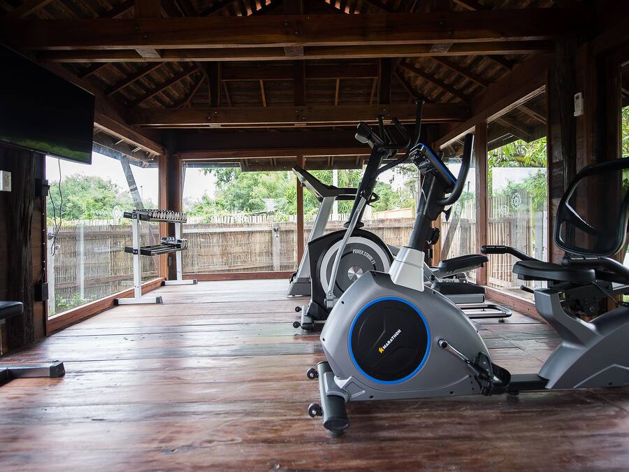 A covered outdoor gym area with exercise equipment, wooden flooring, and a view of a fence and greenery.