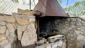 Ein steinerner Grill für den Außenbereich mit einem Metallschornstein, umgeben von einer Steinmauer und einem Maschendrahtzaun.