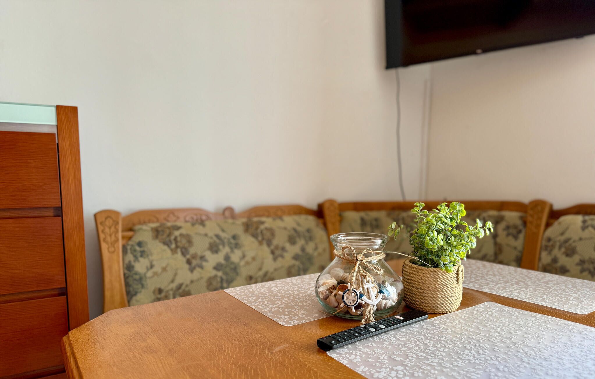 A wooden table with a remote control, a glass jar with seashells and a toy anchor, and a small potted plant on a floral-patterned cushion.