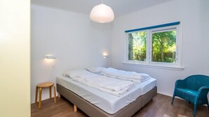A bedroom with a bed, a small stool, a chair, a window with a view of trees, and a pendant light.