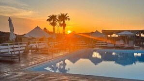 Eine Holzterrasse mit Pool, Palmen und weißen Sonnenschirmen bei Sonnenuntergang.