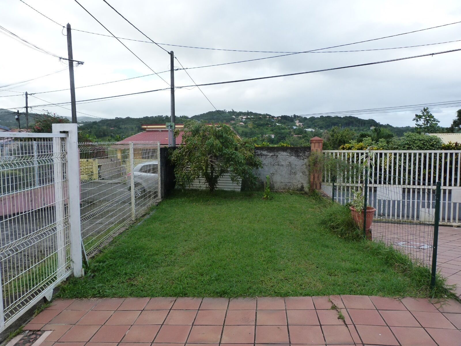 A fenced outdoor area with a grassy lawn, a brick pathway, and a view of a red-roofed building in the background.
