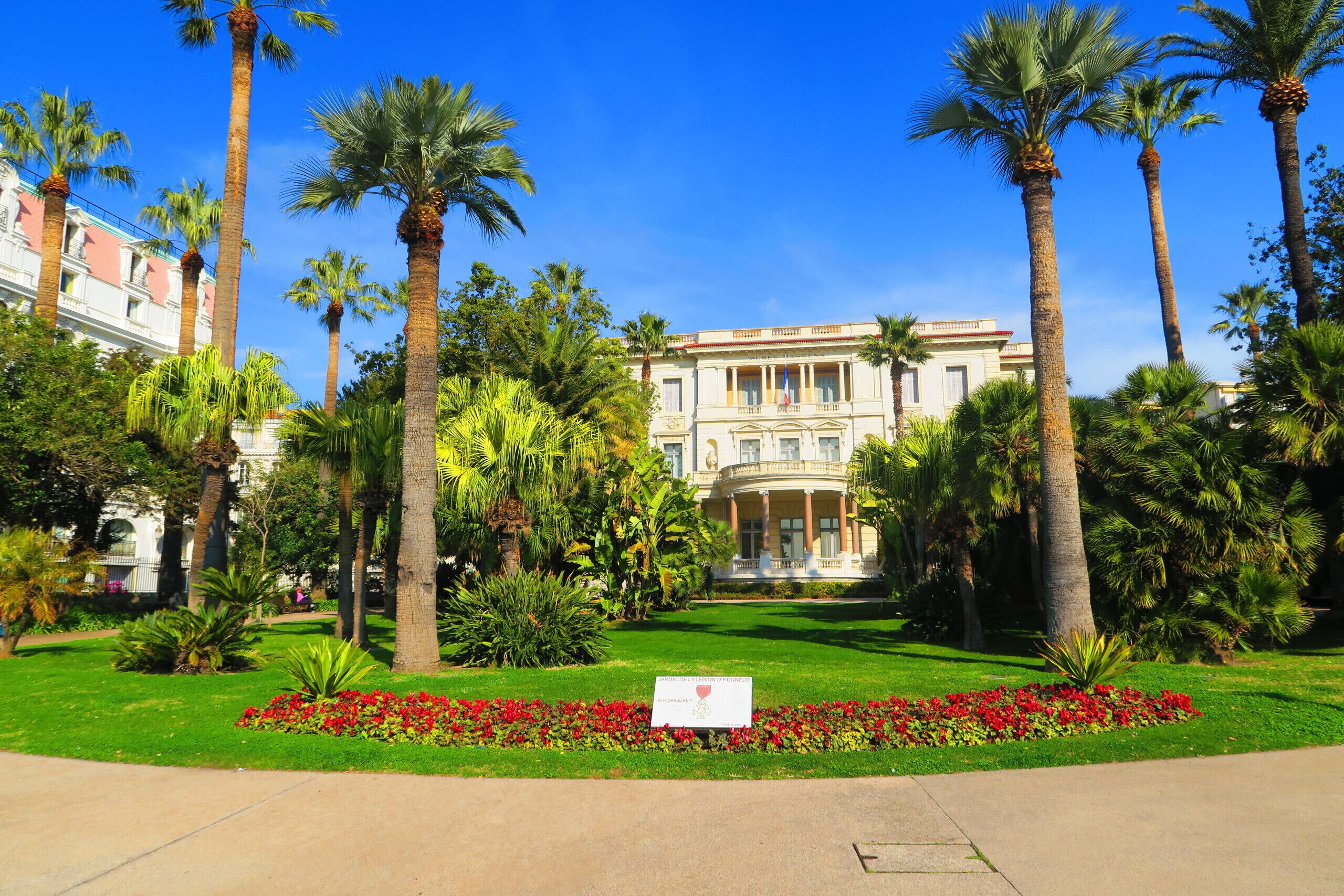 A large, classical building with a symmetrical facade, surrounded by a well-maintained garden with palm trees and red flowers.