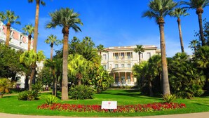 Ein großes, klassisches Gebäude mit symmetrischer Fassade, umgeben von einem gepflegten Garten mit Palmen und roten Blüten.