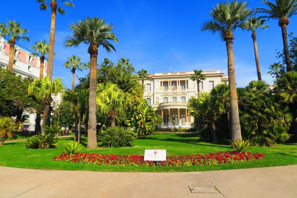 A large, classical building with a symmetrical facade, surrounded by a well-maintained garden with palm trees and red flowers.