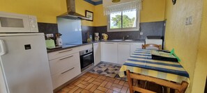 Une cuisine de taille réduite avec des meubles blancs, un four à micro-ondes, un four traditionnel et un évier. On y trouve une fenêtre ornée d’un rideau en dentelle, un sol carrelé et une table recouverte d’une nappe à rayures.