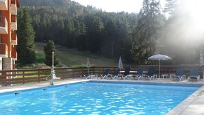 Una piscina con lettini e ombrelloni, circondata da un recinto in legno e un pendio boscoso.