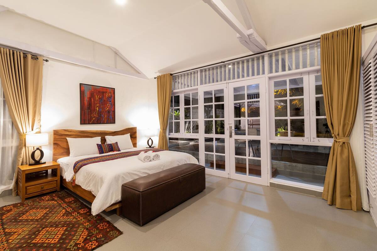 Un dormitorio con una cama grande, cabecera de madera, mesita de noche con lámpara, banca de cuero marrón, alfombra estampada y un ventanal con cortinas.