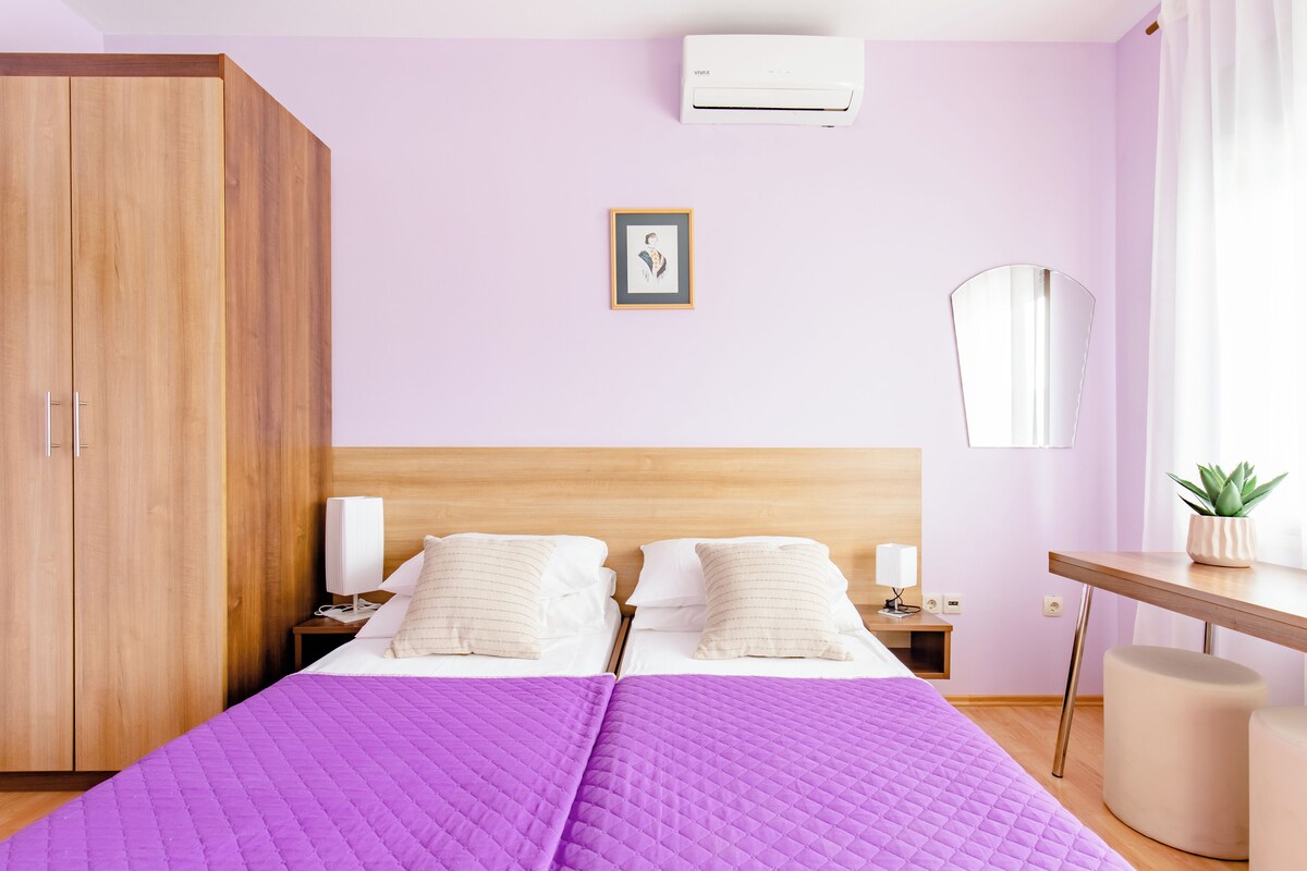 A bedroom with a wooden wardrobe, a bed with a purple quilt, a bedside table with a lamp, and a small plant on a desk.