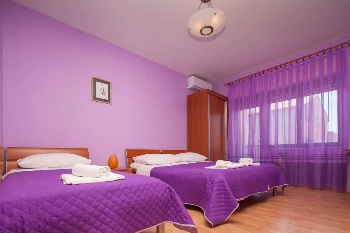 Een kamer met twee bedden, paars beddengoed, een houten garderobe, een lijst met een foto aan de muur, en een raam met paarse gordijnen.