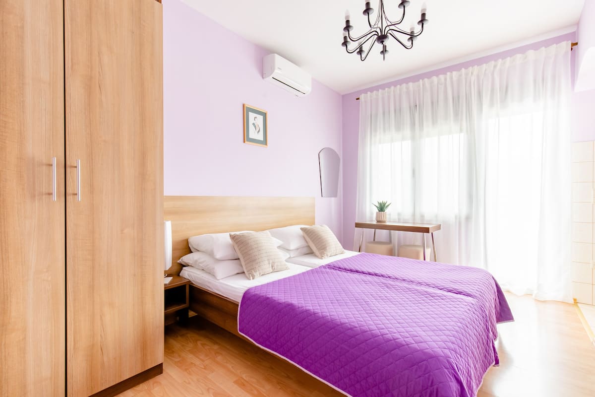 Een slaapkamer met een houten garderobe, een bed met een paars quilt, een nachtkastje met een lamp en een raam met dunne gordijnen.