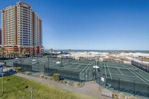 A multi-story hotel with a tennis court in the foreground, surrounded by a sandy beach and ocean view.