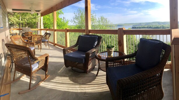 A covered outdoor deck with wicker furniture, a grill, and a view of a lake and forest.