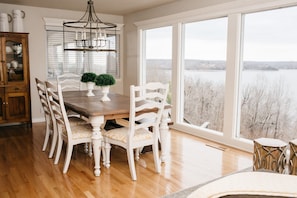A dining area with a wooden table and white chairs, a chandelier, and a view of a lake through large windows.