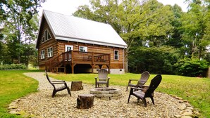 A log cabin with a deck, surrounded by a fire pit and Adirondack chairs.