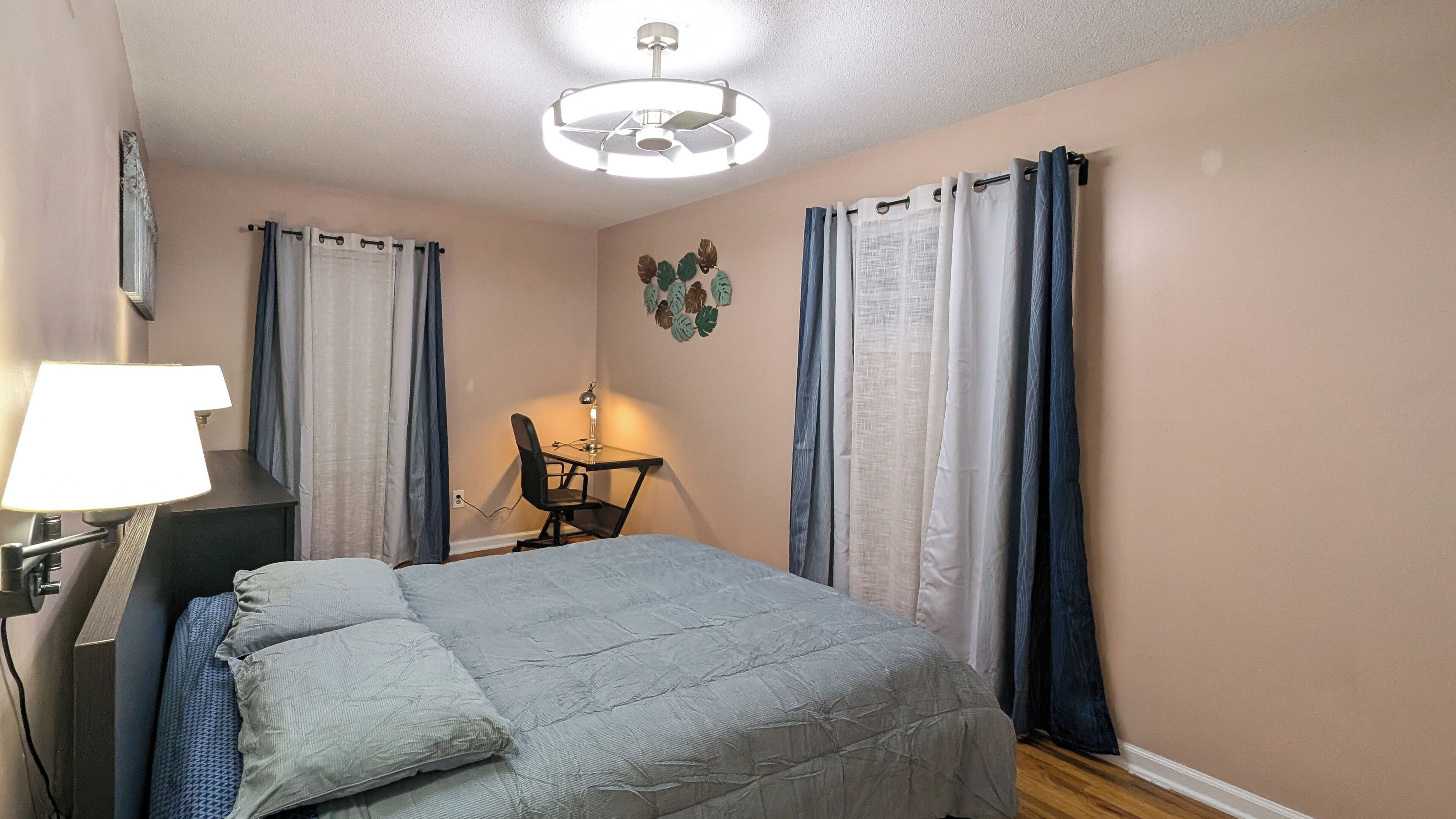 Un dormitorio con cama, escritorio, silla, lámpara y ventilador de techo.