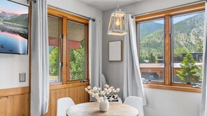 A room with a round wooden table, white chairs, a vase with flowers, and large windows with curtains.