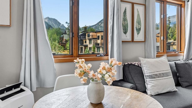A modern living room with a large window, a round table with a vase of flowers, and a sofa with cushions.