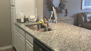 Uma cozinha com bancadas de granito, armários brancos, pia de aço inoxidável e máquina de lavar louça. Há um pássaro decorativo no armário, um relógio de parede e um espelho com design em forma de sol.