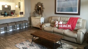 Uma sala de estar com um sofá bege, uma mesa de centro de madeira e um tapete estampado. Há uma área de cozinha com um banquinho de bar e uma placa com a inscrição 'Welcome to the Beach'.