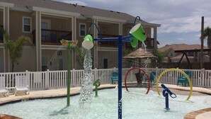 Piscina com recursos aquáticos, incluindo jato d'água e fonte, cercada por uma grade branca e espreguiçadeiras.