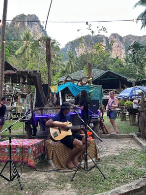 Ein Musiker spielt Gitarre auf einer improvisierten Bühne, mit einem Mikrofonständer, in einer tropischen Umgebung im Freien.