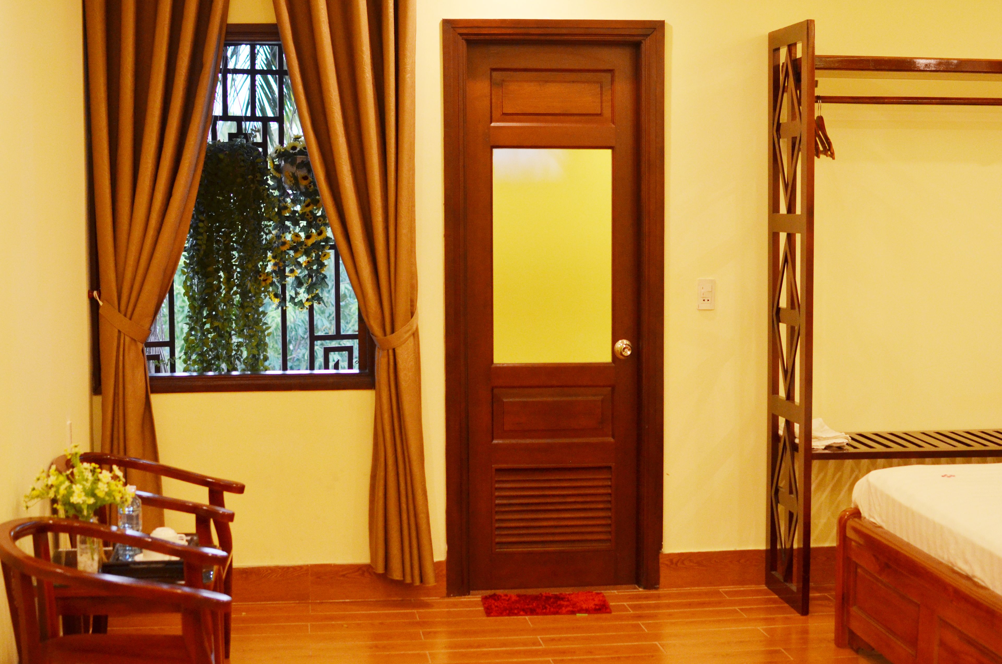 Una habitación con puerta de madera, ventana con cortinas, cama, silla y una mesita con un florero.