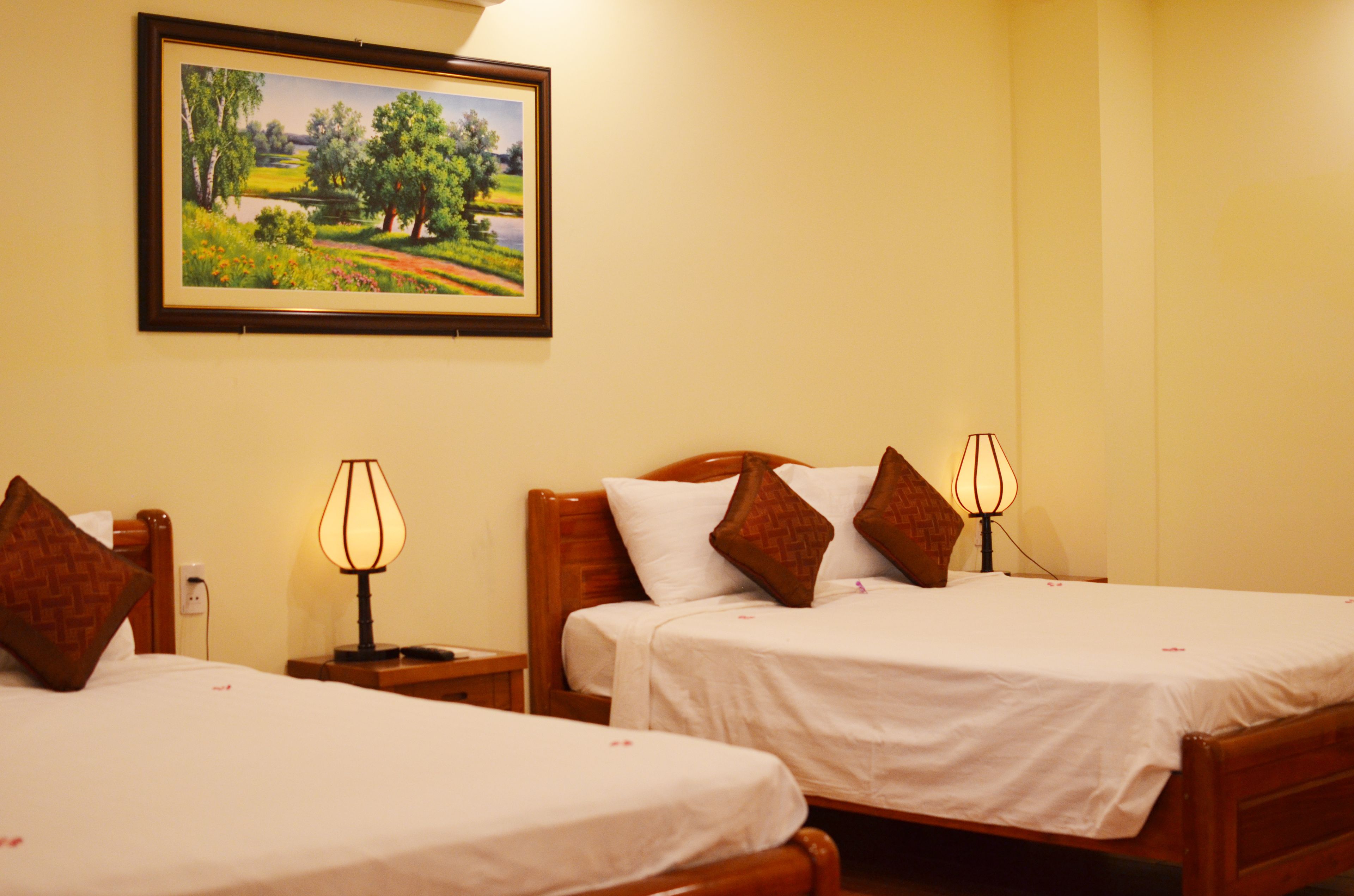 A hotel room with two single beds, each with a wooden headboard, white bedding, and a lamp on the nightstand. A framed landscape painting is on the wall above the beds.