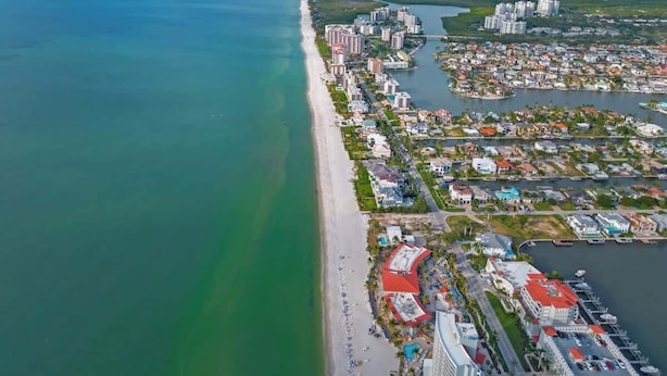 Vista aérea de una ciudad costera con una extensa playa, zona residencial densa y un puerto deportivo.